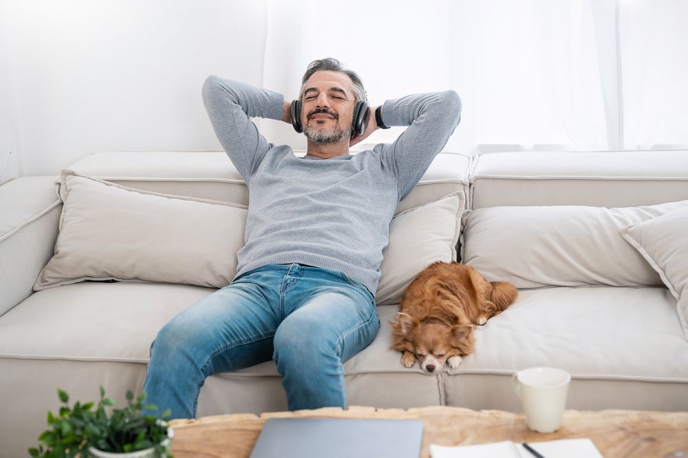 Mann entspannt auf der Couch, Augen zu, hört Musik, Hund schläft neben ihm